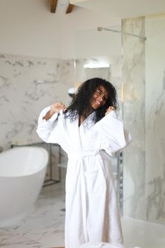 Young pretty black girl wearing white bathrobe and dancing in hotel bathroom with marble walls. Concept of morning relax.