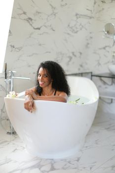 Afro american curly haired nice woman sittling in bath and wearing black swimsuit, marble wall in background. Cocept of bathroom photo shoot and morning hygiene.