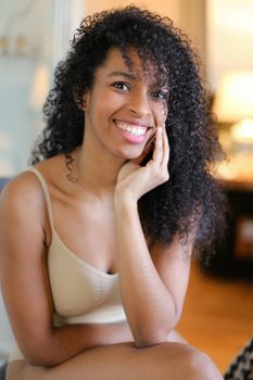 Portrait of young smiling afro american woman sitting in room and wearing beige swimsuit. Concept of hotel photo session in lingerie.