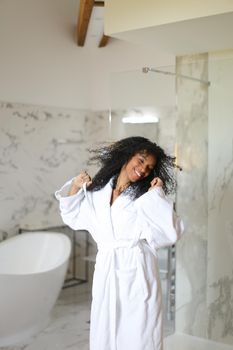 Young black girl wearing white bathrobe and dancing in hotel bathroom with marble walls. Concept of morning relax.