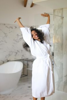 Young happy afro american woman wearing white bathrobe and dancing in hotel bathroom with marble walls. Concept of morning relax.