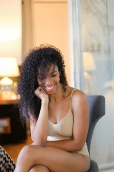 Young happy afro american woman sitting in room and wearing beige swimsuit. Concept of hotel photo session in lingerie.