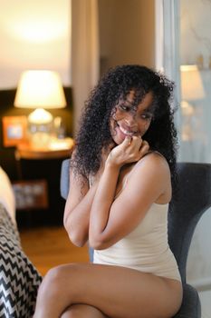Young afro american pretty girl sitting in room and wearing beige swimsuit. Concept of hotel photo session in lingerie.