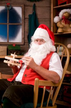 Santa Claus Sitting is a Rocking Chair in His Workshop Painting Toy Airplane. Vertical Composition.