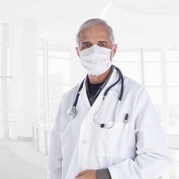 Doctor wearing lab coat and surgical mask with a stethoscope draped around his neck, in high key modern office setting.