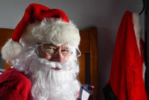 Santa Claus sitting on his bed in his long johns with his Red Suit hanging from a hook in the background.