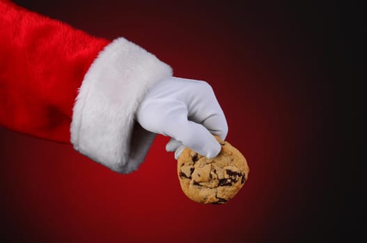 Santa Claus hand holding a chocolate chip cookie over a light to dark red background. Horizontal format showing only hand and arm.
