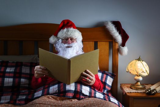 Santa Claus sitting in bed reading a large book before he goes to sleep on Christmas Eve.