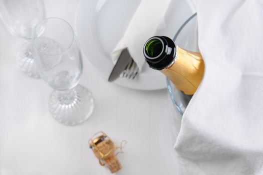 An open champagne bottle in an ice bucket on a table set for dinner. Shallow depth of field, only the champagne bottle is in focus.