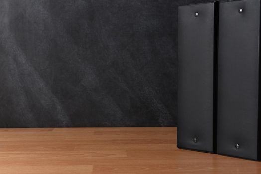 Two black binders standing on a teachers desk in front of a black board.