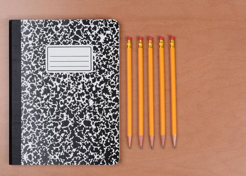 Five sharpened pencils next to a closed theme book on a wood classroom desk. Horizontal format shot form a high angle. 