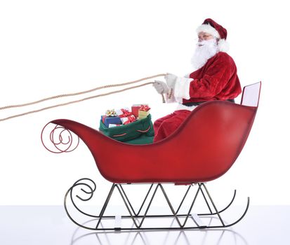 Santa Claus sitting in hi sleigh holidng onto the reins, with a sack of presents at his feet. Isolated on white. 