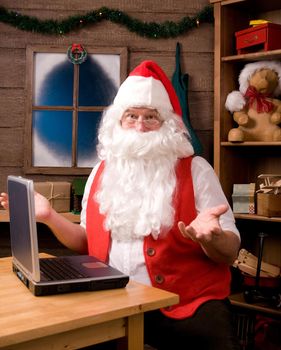 Santa Claus in his workshop with laptop and surrounded by toys and presents. Santa is gesturing with both hands with surprised expression.
