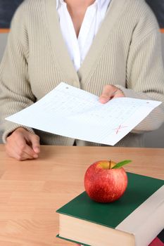 Closeup of an apple atop books on a teachers desk as she hands back graded papers. Shallow depth of field with focus on the foreground.Teacher is unrecognizable and out of focus.