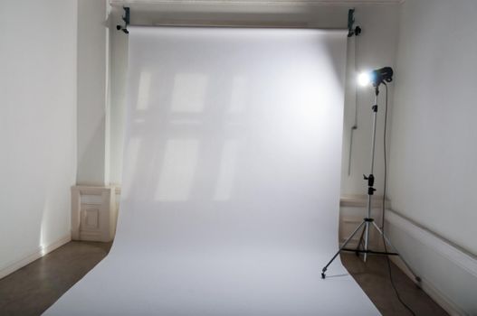 An empty professional photo studio with clean and simple photographic equipment setup of white, plain paper backdrop and a brightly lit flash spotlight on a lamp tripod in a dimmed atelier.