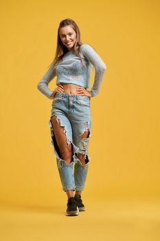 Front view of smiling girl in motion with long brown hair keeping hands on waist. Girl wearing short top with long sleeves, ripped jeans on isolated yellow background. Concept of style, fashion