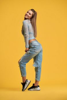 Side view of girl with brown hair tilting head back and pulling toe forward. Lady looking back wearing short top, ripped jeans on isolated yellow background. Concept of style, fashion