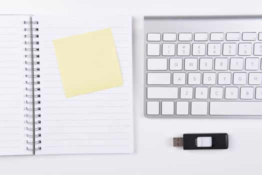 Business Desk: Overhead view of a  notebook with a yellow post-it note, accompanied by a silver computer keyboard with a thumb drive.