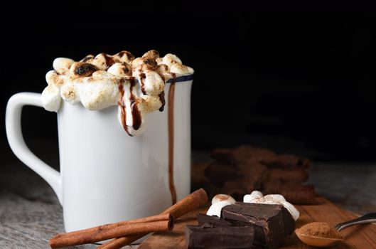 Closeup of a mug of hot cocoa with toasted marshmallows with cookies, chocolate chunks, cinnamon sticks, against a dark background.