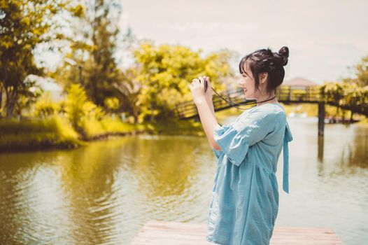 Asian woman in blue dress in public park carrying digital mirrorless camera and taking photo without facial mask in happy mood. People lifestyle and leisure concept. Outdoor travel and Nature theme.