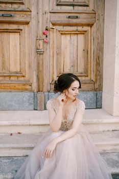 Bride sits on the steps of an ancient building in front of a wooden door. High quality photo