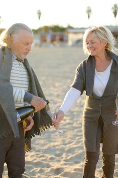 Focus on pouring champagne by elderly husband with caucasian wife on sand beach. Concept of picnic in open air.