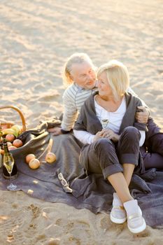 Senior caucasian husband hugging elderly wife sitting on plaid with champagne on sand beach. Concept of pensioners on picnic.