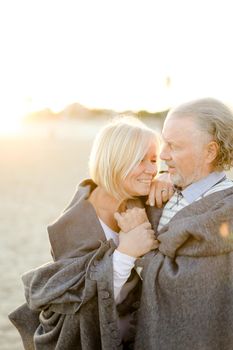 Sunshine photo of senior husband hugging blonde wife wearing plaid on sand beach. Concept of happy elderly couple and relationship.