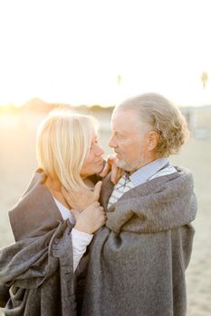Sunshine photo of caucasian senior husband hugging blonde wife wearing plaid on sand beach. Concept of happy elderly couple and relationship.