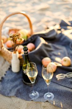 Focus on bottle of champagne and glasses, basket with fruits on plaid and sand beach in blurry background. Concept of picnic composition.
