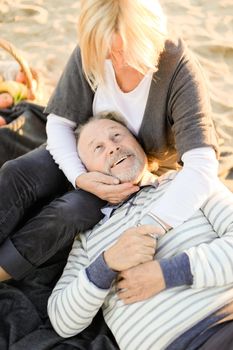 Senior husband lying on blonde wife knees, sand beach in background. Concept of pensioners on picnic and happy elderly couple.