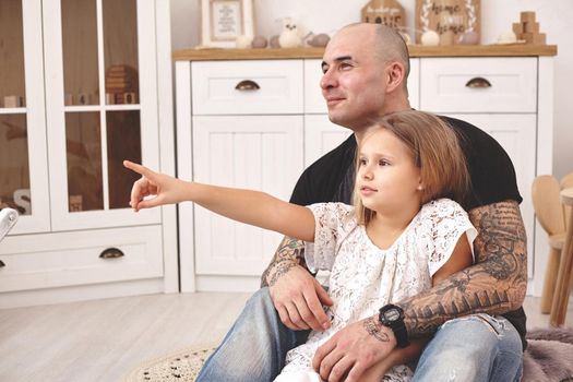 White modern kid's room whith a wooden furniture. Adorable daughter with a long blond hair wearing a white dress. Daddy with tattoos is hugging her while she is pointing on something, they are looking away. Friendly family spending their free time together sitting on a pillows.