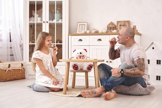 White modern kid's room whith a wooden furniture. Adorable daughter wearing a white dress is looking with tenderness at her loving father. They are drinking tea from a toy dishes in a modern kid's room whith a wooden furniture. Daddy with tattoos is laughing and looking at her. Friendly family spending their free time together sitting on a pillows.