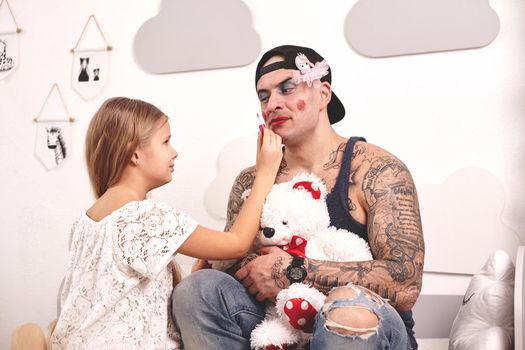 Funny time Tattoed father in a cap and his child are playing at home. Pretty girl in a white dress is putting a rouge at her dady's cheeks while he is holding a toy bear, in her bedroom. Family holiday and togetherness.