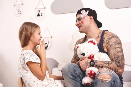 Funny time Tattoed father in a cap, whith a toy bear in his hands, and his child are playing at home. Cute girl in a white dress is doing makeup to her dad and laughing at him, in her bedroom. Family holiday and togetherness.
