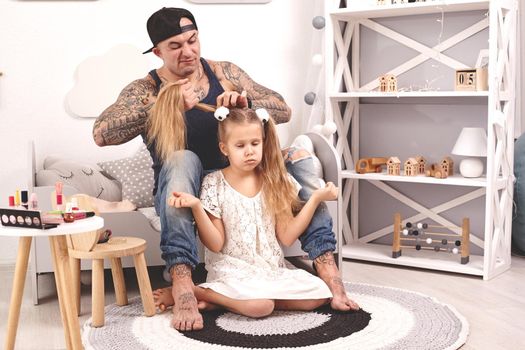 Funny time Tattoed father in a cap and his child are playing at home. Young dad is doing his daughter's hair while she is sitting on a rug in her bedroom. Family holiday and togetherness.