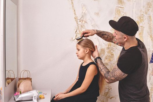 Cute little daughter and her tattoed dad are playing together near a mirror. Handsome dad is doing his daughter's hair. Family holiday and togetherness.