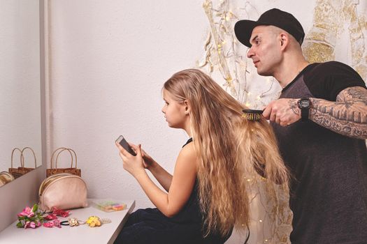 Cute little daughter and her tattoed dad are playing together near a mirror. Handsome dad is drying his daughter's hair while she is looking at her phone. Family holiday and togetherness.