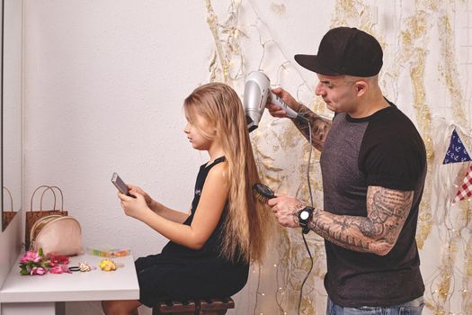 Cute little daughter and her tattoed dad are playing together near a mirror. Dad is drying his daughter's hair while she is looking at her phone. Family holiday and togetherness.
