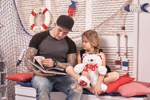 Handsome tattoed man in a cap is spending time with his little cute daughter. They are sitting on a white bench with some red pillows, in a room decorated in a marine style. She is holding a toy bear. Reading fairytales while daughter is sitting nearby. Happy family.