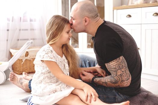 White modern kid's room whith a wooden furniture. Adorable daughter with a blond long hair wearing a white dress. Daddy with tattoos is kissing her while holding her hand. Friendly family spending their free time together sitting on a pillows.