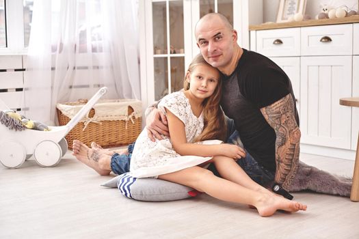 White modern kid's room whith a wooden furniture. Adorable daughter whith a long blond hair wearing a white dress . Daddy with tattoos is hugging her and they are looking at the camera. Friendly family spending their free time together sitting on a pillows.