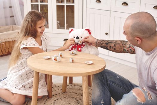 White modern kid's room whith a wooden furniture. Adorable daughter whith a long blond hair wearing a white dress. They are treating a toy bear with tea from a toy dishes in a modern kid's room whith a wooden furniture. Friendly family spending their free time together sitting on a pillows.