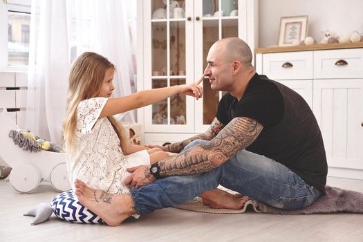 White modern kid's room whith a wooden furniture. Adorable daughter wearing a white dress is touching daddy's nose and looking with tenderness at him. Daddy with tattoos is hugging her. Friendly family spending their free time together sitting on a pillows.