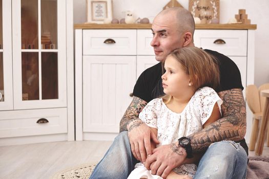 White modern kid's room whith a wooden furniture. Adorable daughter with a long blond hair wearing a white dress. Daddy with tattoos is hugging her and they are looking somewhere. Friendly family spending their free time together sitting on a pillows.