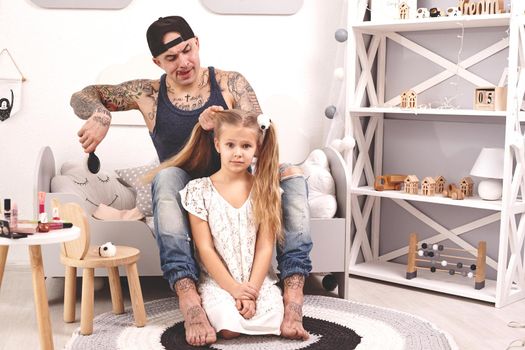 Funny time Attractive tattoed father in a cap and his child are playing at home. Dad is doing his daughter's hair while she is sitting on a rug in her bedroom. Family holiday and togetherness.