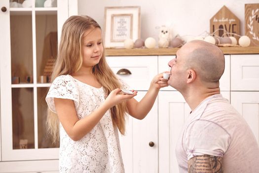 White modern kid's room whith a wooden furniture. Adorable daughter whith a long blond hair wearing a white dress is treating her daddy with some tea. They are drinking it from a toy dishes in a modern kid's room whith a wooden furniture. Daddy with tattoos is trying a tea. Friendly family spending their free time together sitting on a pillows.