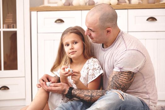 White modern kid's room whith a wooden furniture. Adorable daughter whith a long blond hair wearing a white dress is looking at the camera. They are drinking tea from a toy dishes in a modern kid's room whith a wooden furniture. Daddy with tattoos is hugging and looking at her. Friendly family spending their free time together sitting on a pillows.