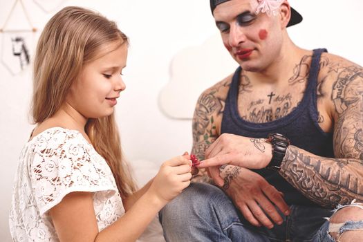 Funny time Tattoed father in a cap and his child are playing at home. Nice girl in a white dress is applying a red nail polish to her dad's fingers in a bedroom. Family holiday and togetherness.