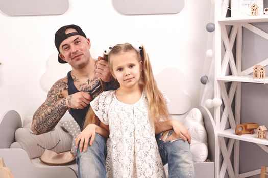 Funny time Tattoed father in a cap and his child are playing at home. Dad is doing his daughter's hair in her bedroom. They are looking happy. Family holiday and togetherness.